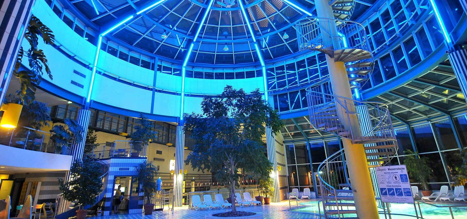 Wunderschöne Nachtaufnahme von der Innenhalle des Wellenspiels. Im Vordergrund führt die Wendeltreppe zur Riesenrutsche hinauf. Die Innenbeleuchtung und viele Pflanzen schaffen eine gemütliche, aber auch magische Atmosphäre.