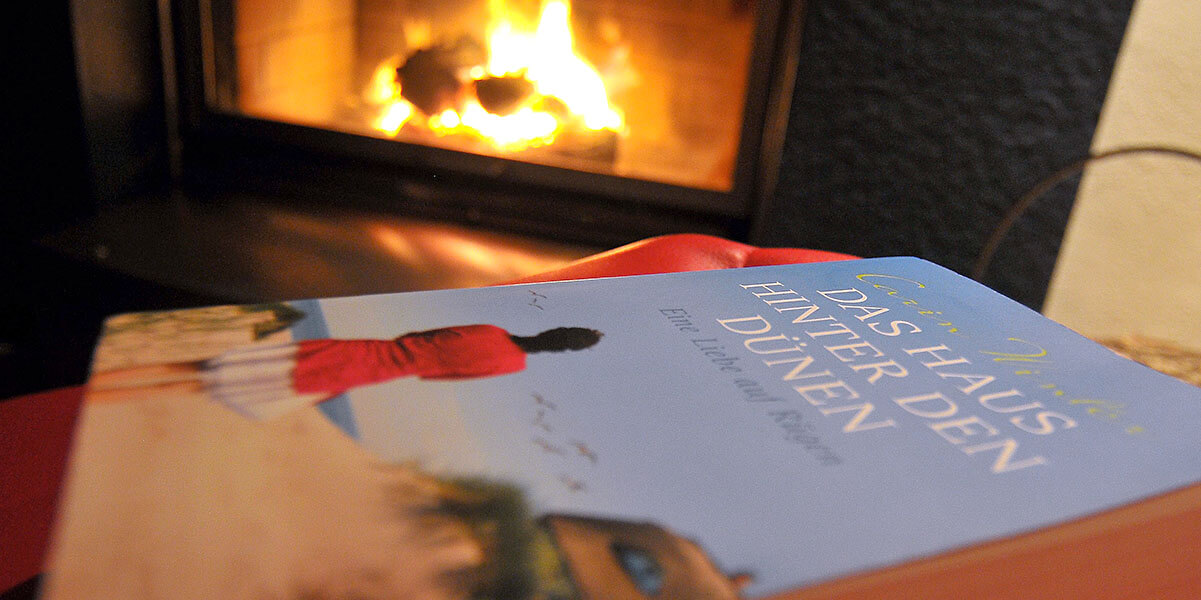 Sauna - Slider - Mitternachtssauna - Im Vordergrund liegt das Buch "Das Haus hinter den Dünen", im Hintergrund brennt das Kaminfeuer